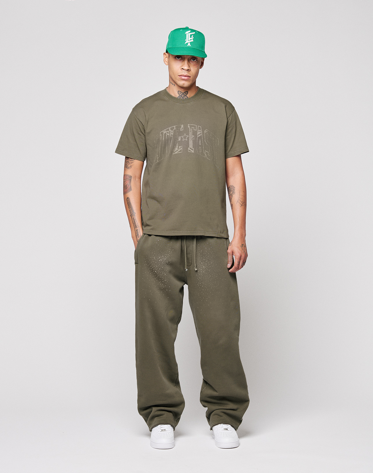 A person stands facing forward wearing the olive green Live Fast Rhinestone Tee by LIVE FAST DIE YOUNG, matching baggy sweatpants, a green baseball cap, and white sneakers against a plain light gray background.