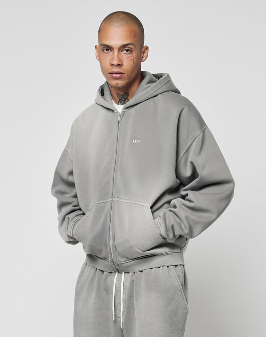 A person with a shaved head and visible tattoos wears the LIVE FAST DIE YOUNG Basic Hooded Zip in light gray with subtle logo print, paired with matching sweatpants, standing against a plain, light background with hands in pockets.