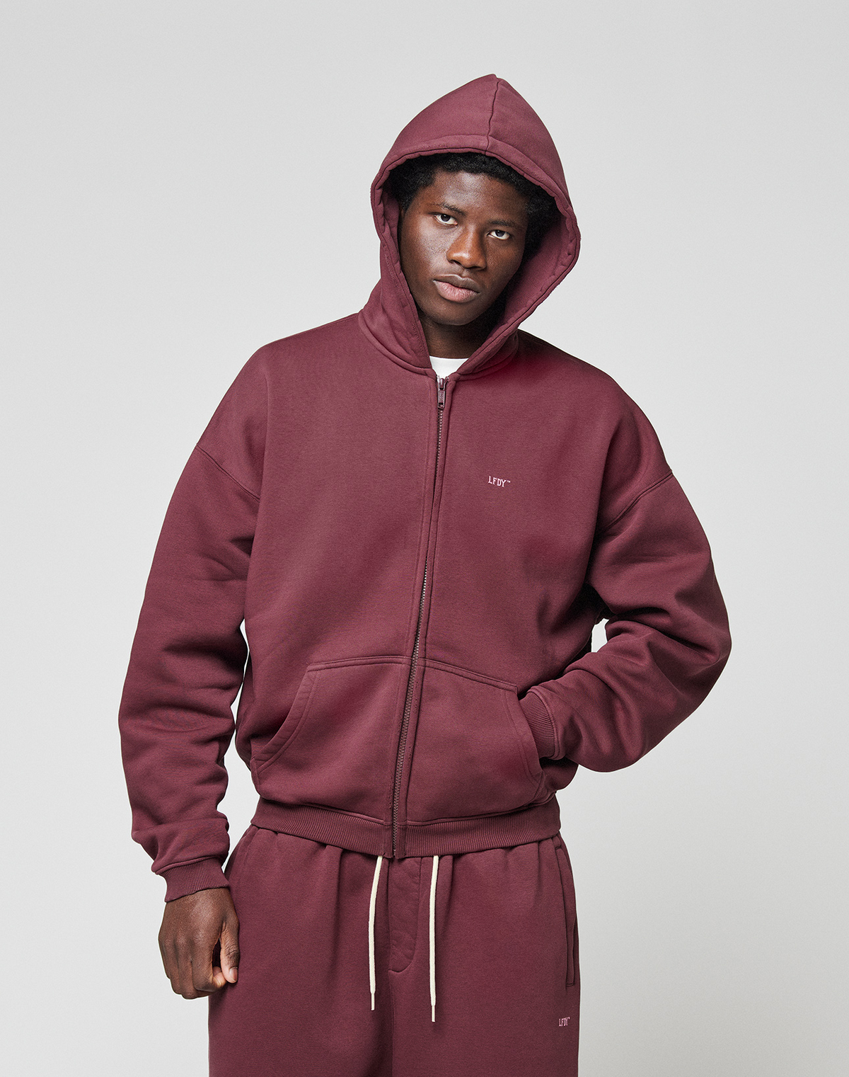 A person wears the LIVE FAST DIE YOUNG Basic Hooded Zip in maroon with the hood up and matching sweatpants, standing against a light gray background and looking at the camera with one hand in their pocket.