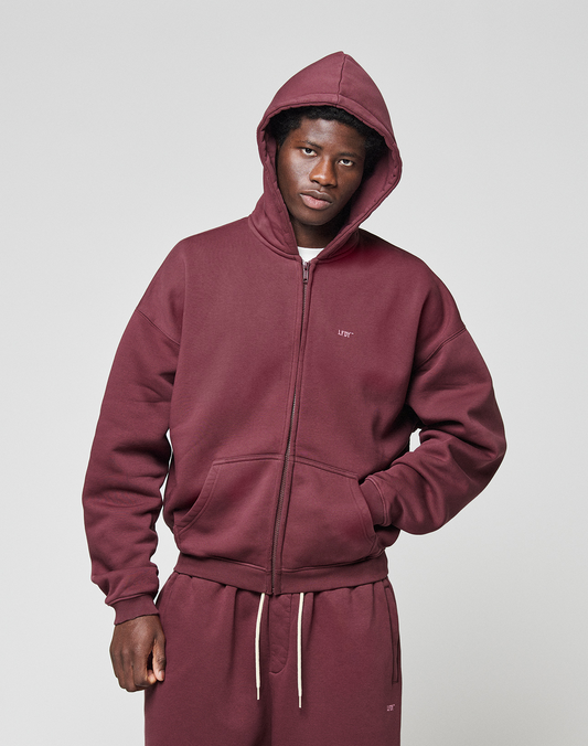 A person wears the LIVE FAST DIE YOUNG Basic Hooded Zip in maroon with the hood up and matching sweatpants, standing against a light gray background and looking at the camera with one hand in their pocket.