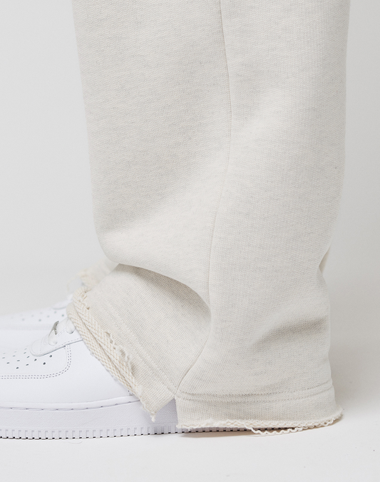 A close-up of a person wearing LIVE FAST DIE YOUNG LFDY Heavyweight Loungepants in off-white with frayed hems and white sneakers, standing against a plain light background. The focus is on the lower leg and foot.