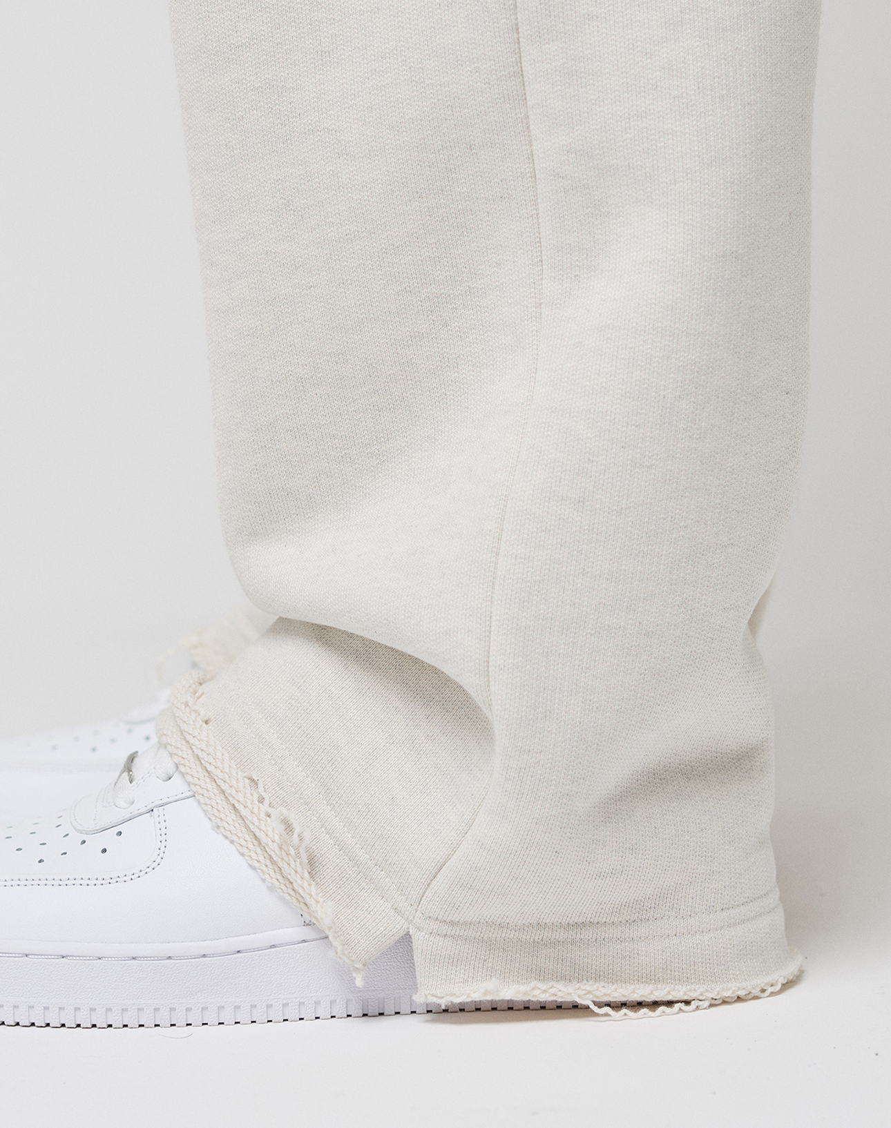 A close-up of a person wearing LIVE FAST DIE YOUNG LFDY Heavyweight Loungepants in off-white with frayed hems and white sneakers, standing against a plain light background. The focus is on the lower leg and foot.