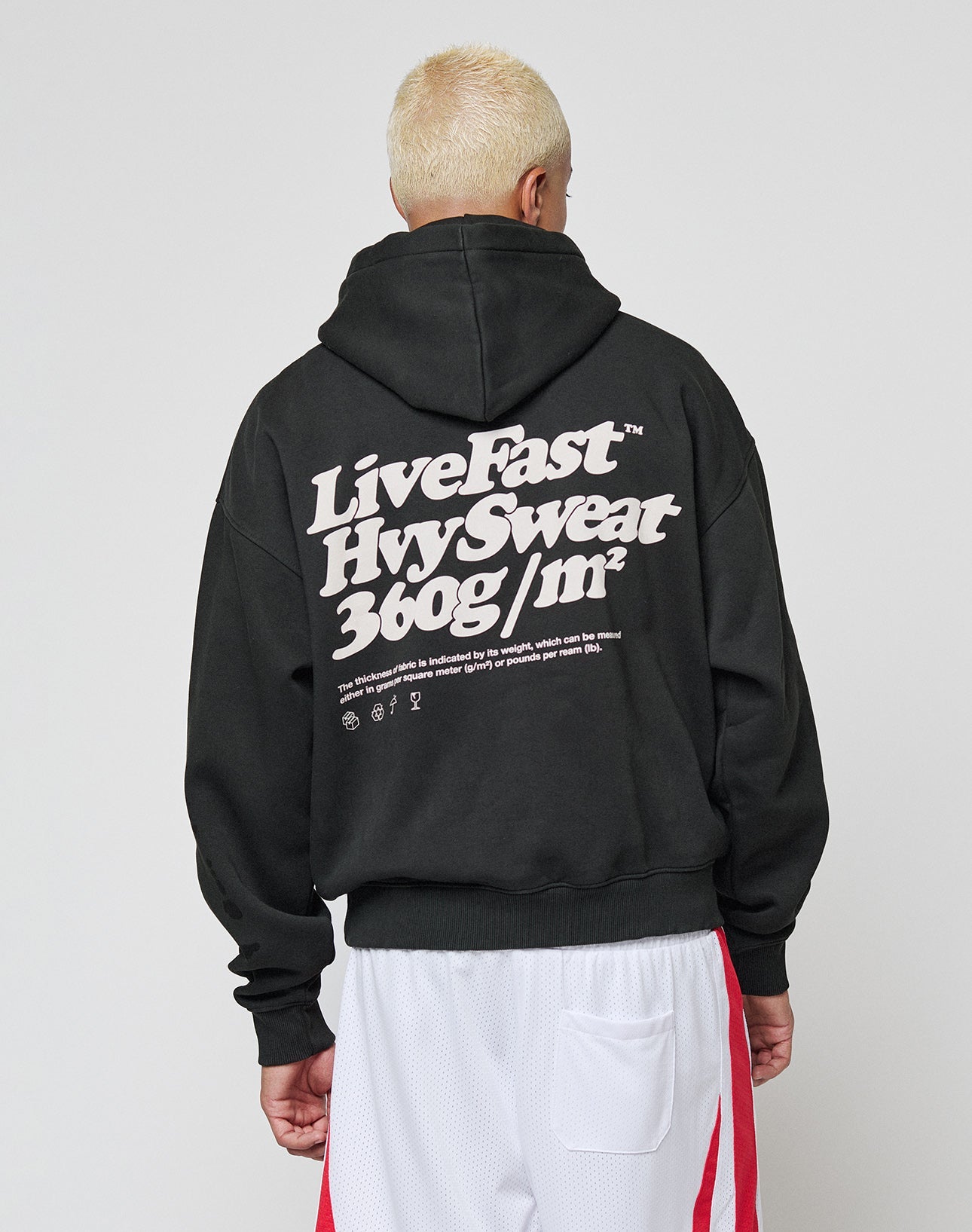 Someone with short, bleached blond hair faces away, wearing a LIVE FAST DIE YOUNG Basic 360 Hooded oversize black hoodie with bold white text and white athletic shorts with red stripes.