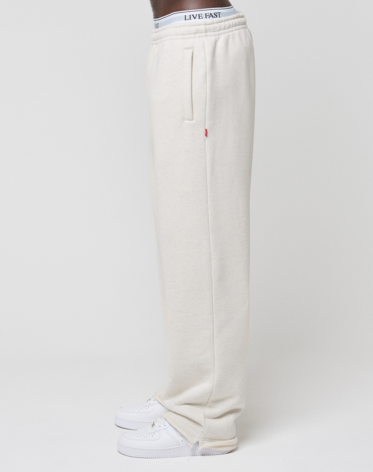 A person stands in profile wearing LIVE FAST DIE YOUNG LFDY Heavyweight Loungepants in off-white 650gsm premium cotton, styled with white sneakers and a waistband showing LIVE FAST text; background is plain white.