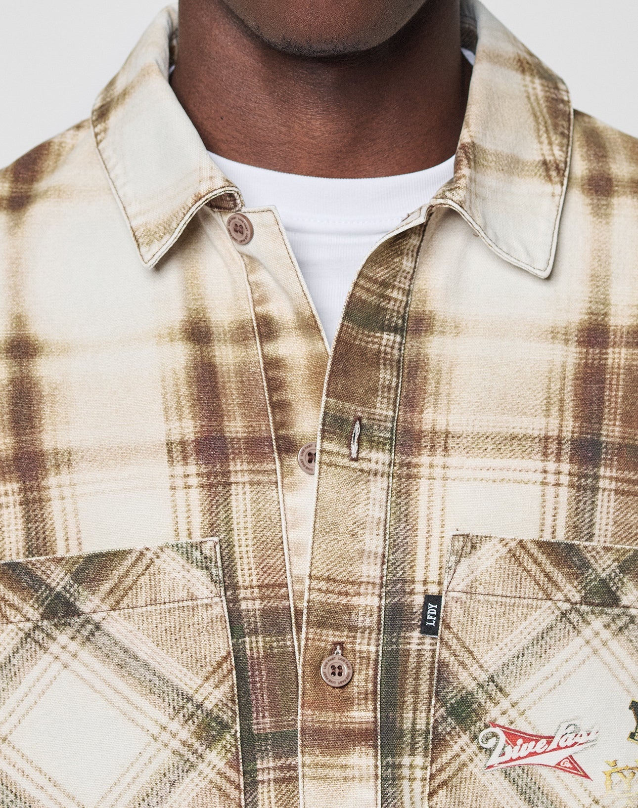 A close-up of someone wearing the LIVE FAST DIE YOUNG Faded Flannel in beige and brown plaid, made from soft cotton. The oversized shirt features two chest pockets and visible stitching, styled over a white T-shirt. The face is mostly out of frame.