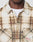 A close-up of someone wearing the LIVE FAST DIE YOUNG Faded Flannel in beige and brown plaid, made from soft cotton. The oversized shirt features two chest pockets and visible stitching, styled over a white T-shirt. The face is mostly out of frame.