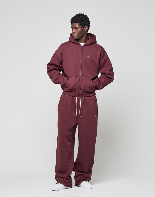 A man stands against a plain light background wearing an oversize maroon LIVE FAST DIE YOUNG Basic Hooded Zip tracksuit with branded zip-up hoodie and drawstring pants, white sneakers, hands in his hoodie pockets, looking to his left.