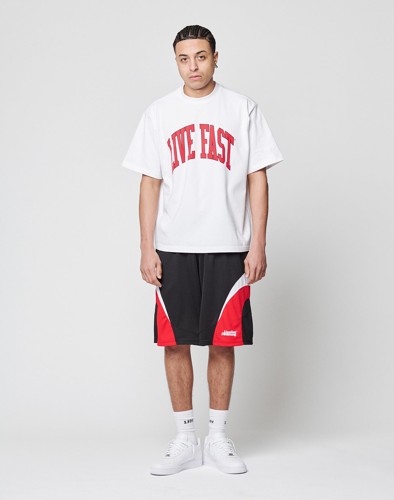 A man with braided hair stands against a plain background, wearing an oversize white LIVE FAST DIE YOUNG Live Fast College Tee with front print, paired with black and red athletic shorts, white socks, and white sneakers.