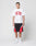 A man with braided hair stands against a plain background, wearing an oversize white LIVE FAST DIE YOUNG Live Fast College Tee with front print, paired with black and red athletic shorts, white socks, and white sneakers.
