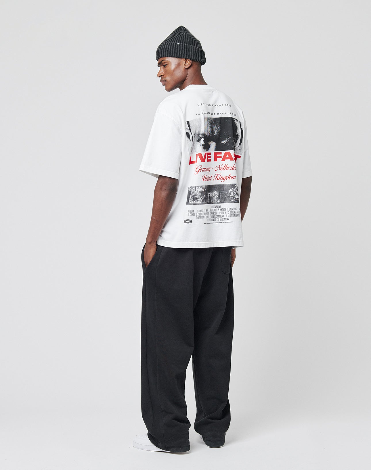 A man in a black beanie, loose black pants, and the LIVE FAST DIE YOUNG LFDY Cinema Tee with front and back graphics stands in profile against a plain white background.