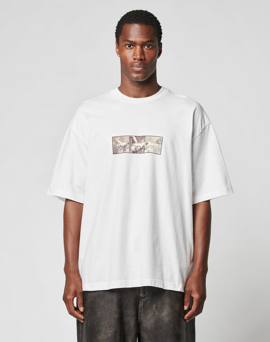 A man with short hair and a neutral expression faces forward against a light gray background, wearing the oversize-fit white LFDY Trinity Tee by LIVE FAST DIE YOUNG with a small reflective chest print.