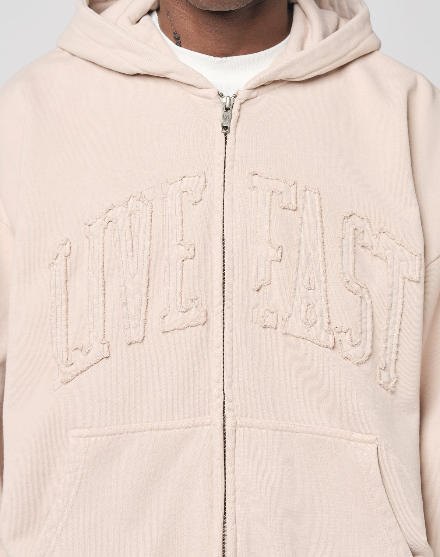 A person wears the LIVE FAST DIE YOUNG Ripped College Hooded Zip hoodie in light beige Baumwolle, featuring bold LIVE FAST embroidery. Only their chest and part of their face are visible.