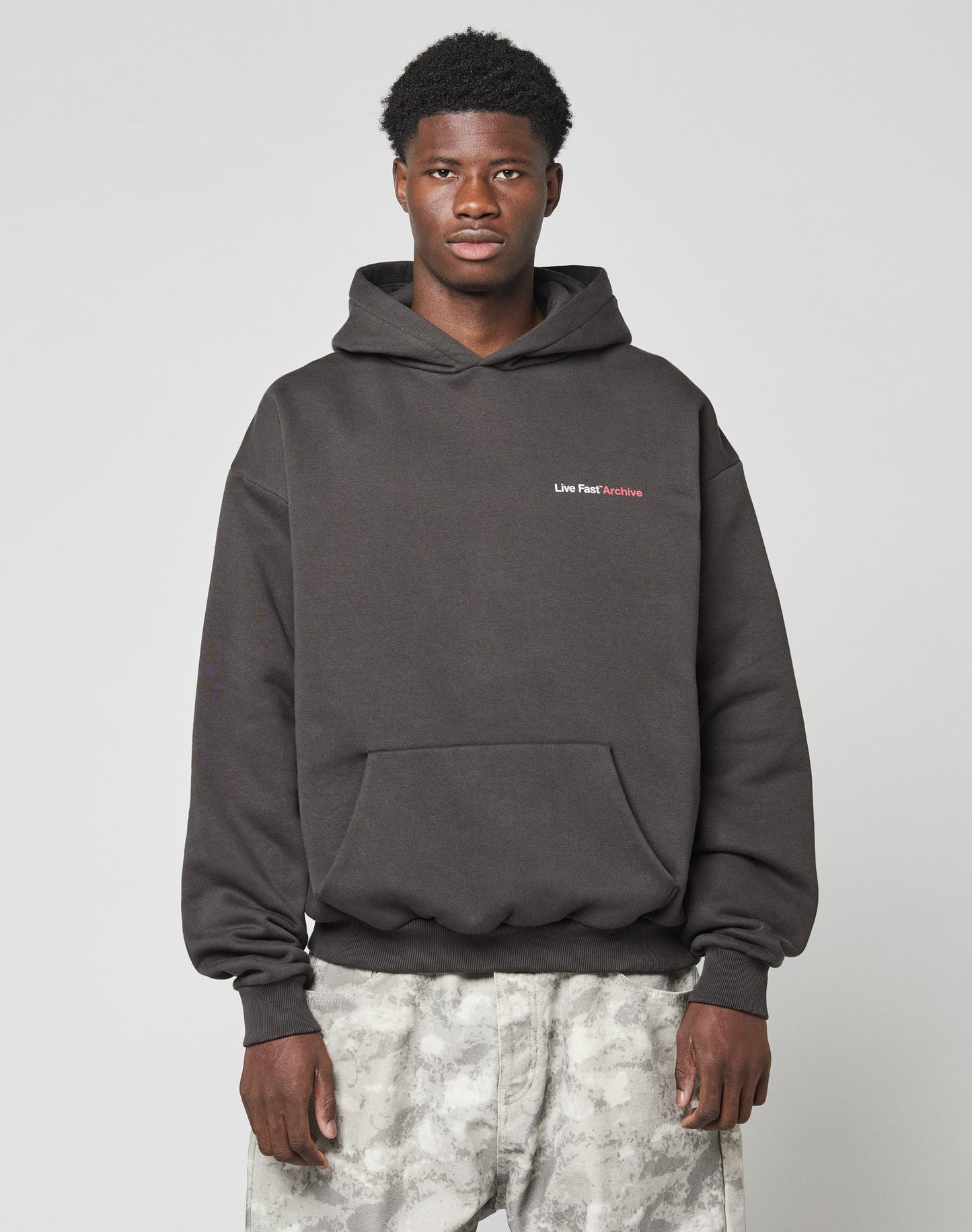 A man stands against a plain light background wearing an oversize fit black LIVE FAST DIE YOUNG LFDY F*ck Phones Hooded sweatshirt with Live Fast Archive text on the left chest and light gray camouflage pants.