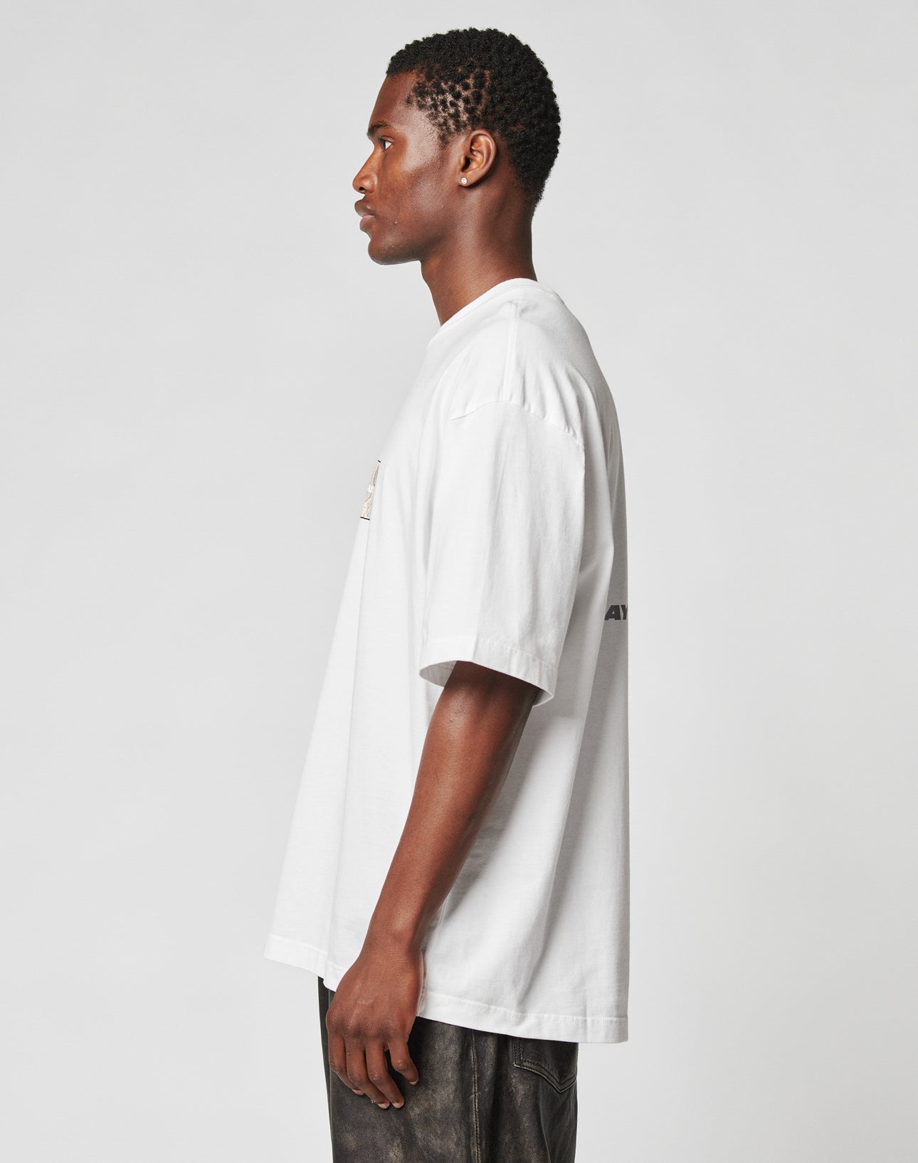 A man in profile faces left, wearing the LFDY Trinity Tee by LIVE FAST DIE YOUNG—an oversize fit white t-shirt made from 100% Baumwolle—with dark pants against a plain light gray background.