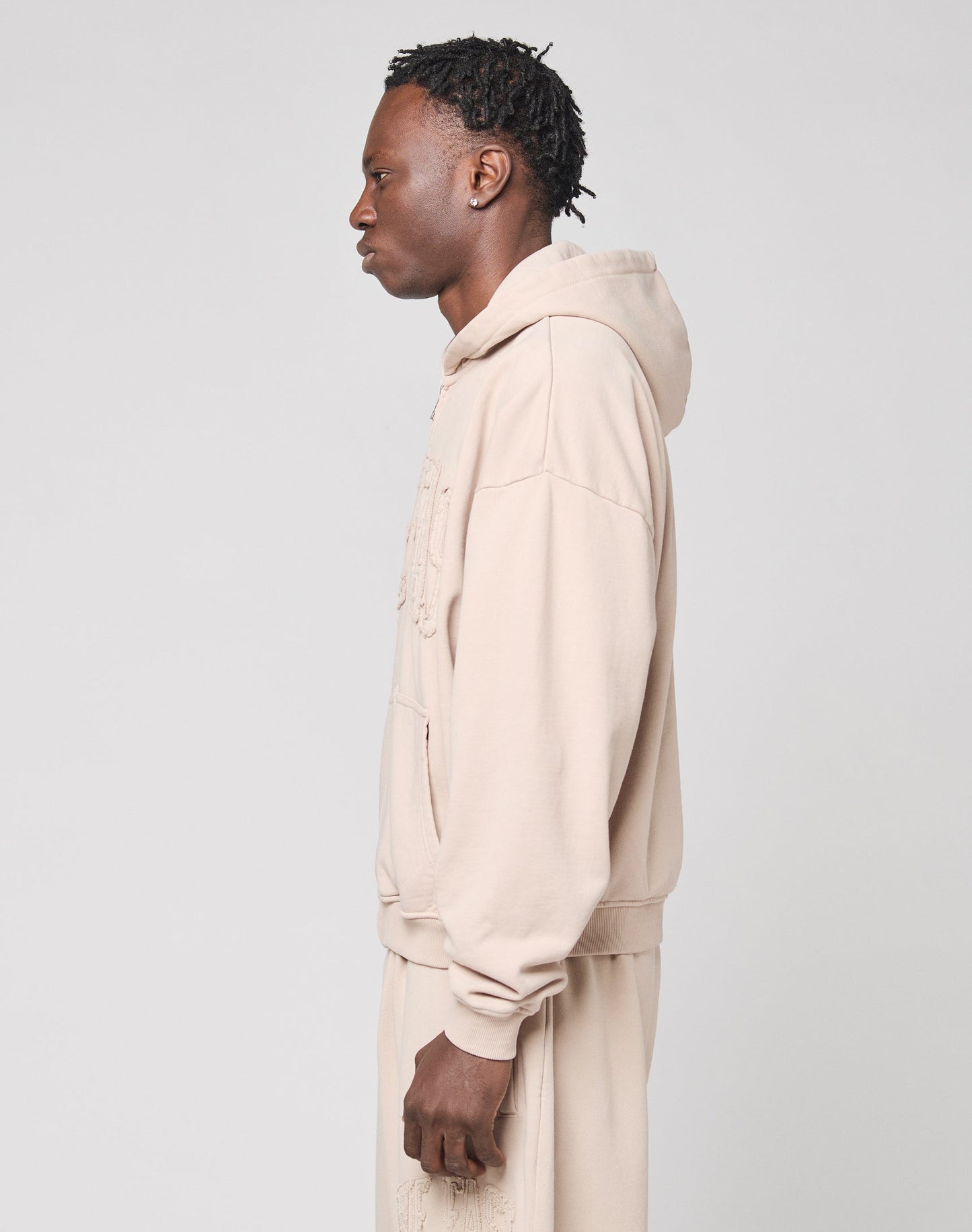 A person with short, braided hair stands in profile against a plain light background, wearing the LIVE FAST DIE YOUNG Ripped College Hooded Zip—an oversized beige sweatshirt made from soft Baumwolle.