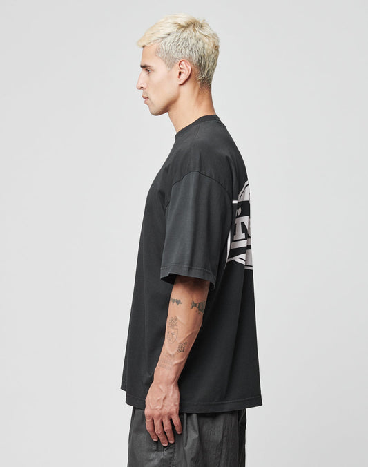 A person with short, bleached blond hair stands in profile, wearing the LIVE FAST DIE YOUNG LFDY Bulge Tee—an oversize black T-shirt with white front and back prints—and dark pants. Tattoos cover their left forearm. The background is plain light gray.