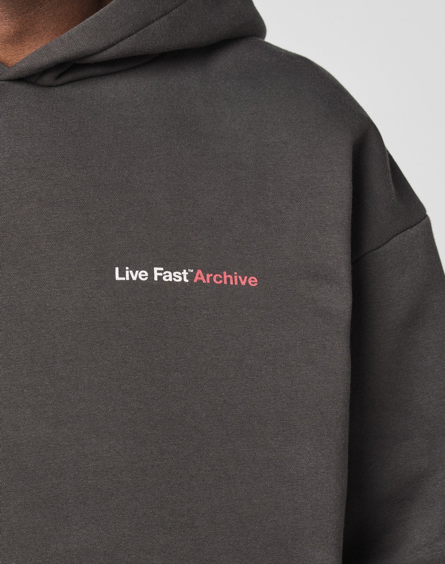 Close-up of the LIVE FAST DIE YOUNG LFDY F*ck Phones Hooded, an oversized dark hoodie with Live Fast Archive printed in small white and pink letters on the chest; upper shoulder and neck of a person are visible.