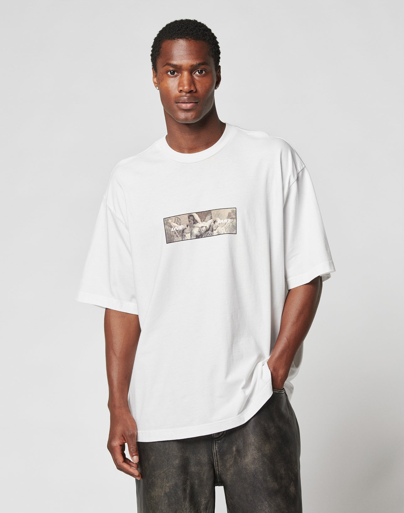 A man wears the LIVE FAST DIE YOUNG LFDY Trinity Tee, a loose white t-shirt featuring a small reflective graphic on the chest, standing with one hand in his pocket against a plain light background.