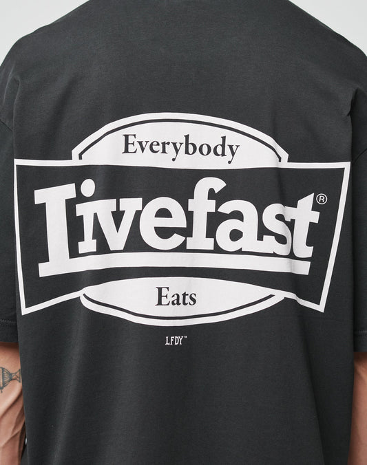 A person wears the LFDY Bulge Tee by LIVE FAST DIE YOUNG, an oversize black cotton T-shirt with a white rectangular graphic on the back that reads Everybody Livefast Eats LFDTM; only the back print and part of their arm are visible.