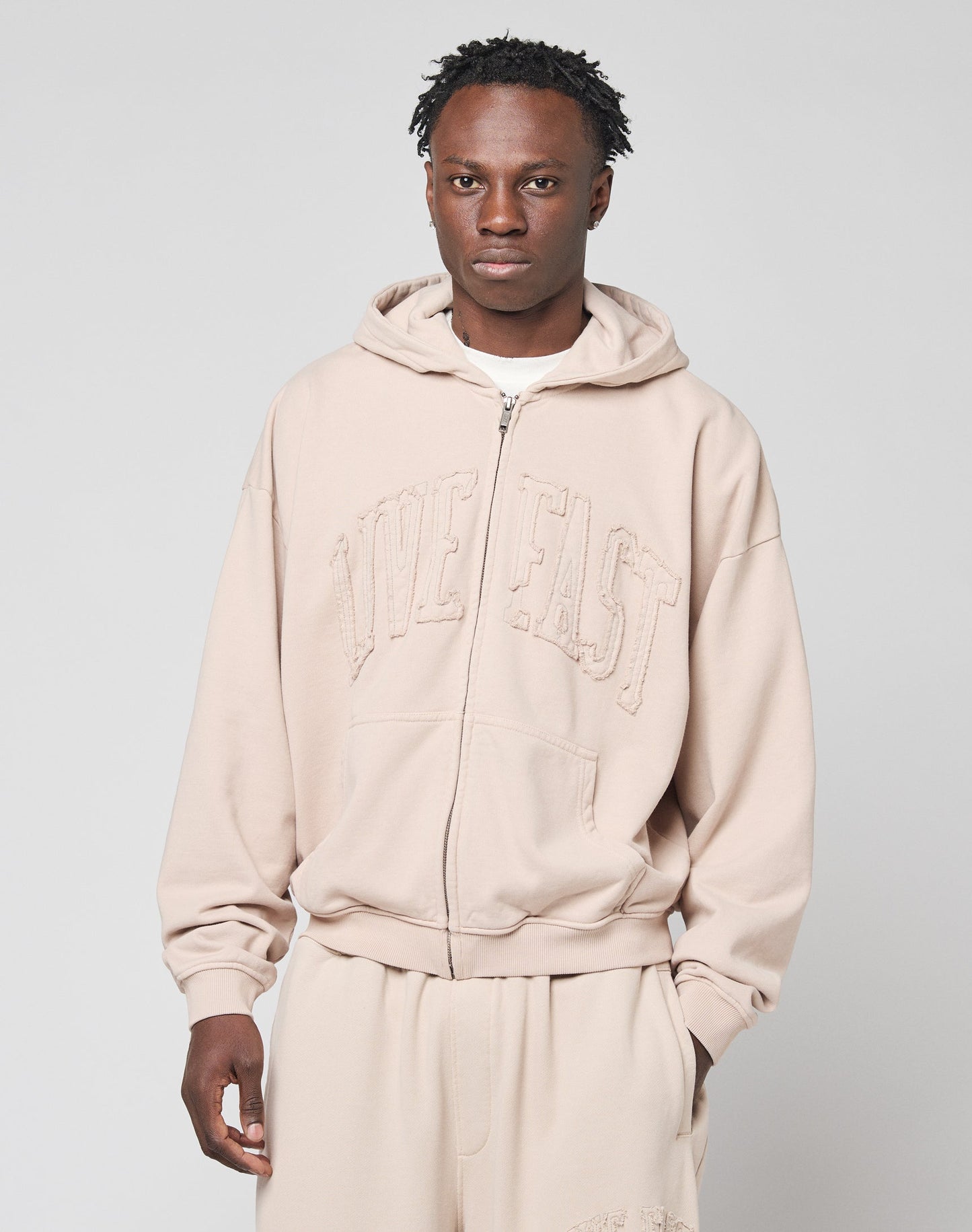 A man wears the LIVE FAST DIE YOUNG Ripped College Hooded Zip in light beige with matching sweatpants, standing against a plain light gray background and looking directly at the camera with a neutral expression.
