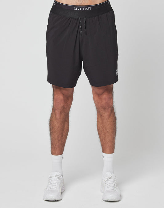 A person stands against a plain white background wearing LIVE FAST DIE YOUNG’s Live Fast Performance Shorts—black with LIVE FAST on the elastic waistband, regular fit—paired with white socks and sneakers. The photo is cropped at the waist and ankles.