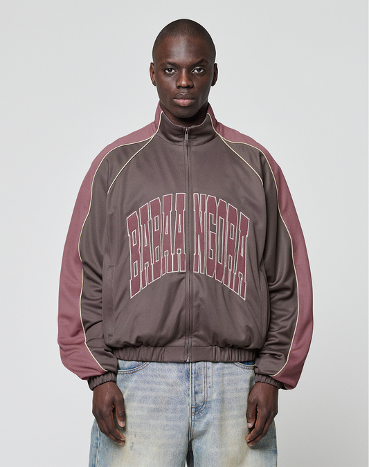 A man with short hair and light blue jeans faces forward, wearing the LIVE FAST DIE YOUNG LFDY x Baba Angora Trackjacket in brown and mauve, featuring bold BABA NIGORA lettering across the chest against a plain background. | model