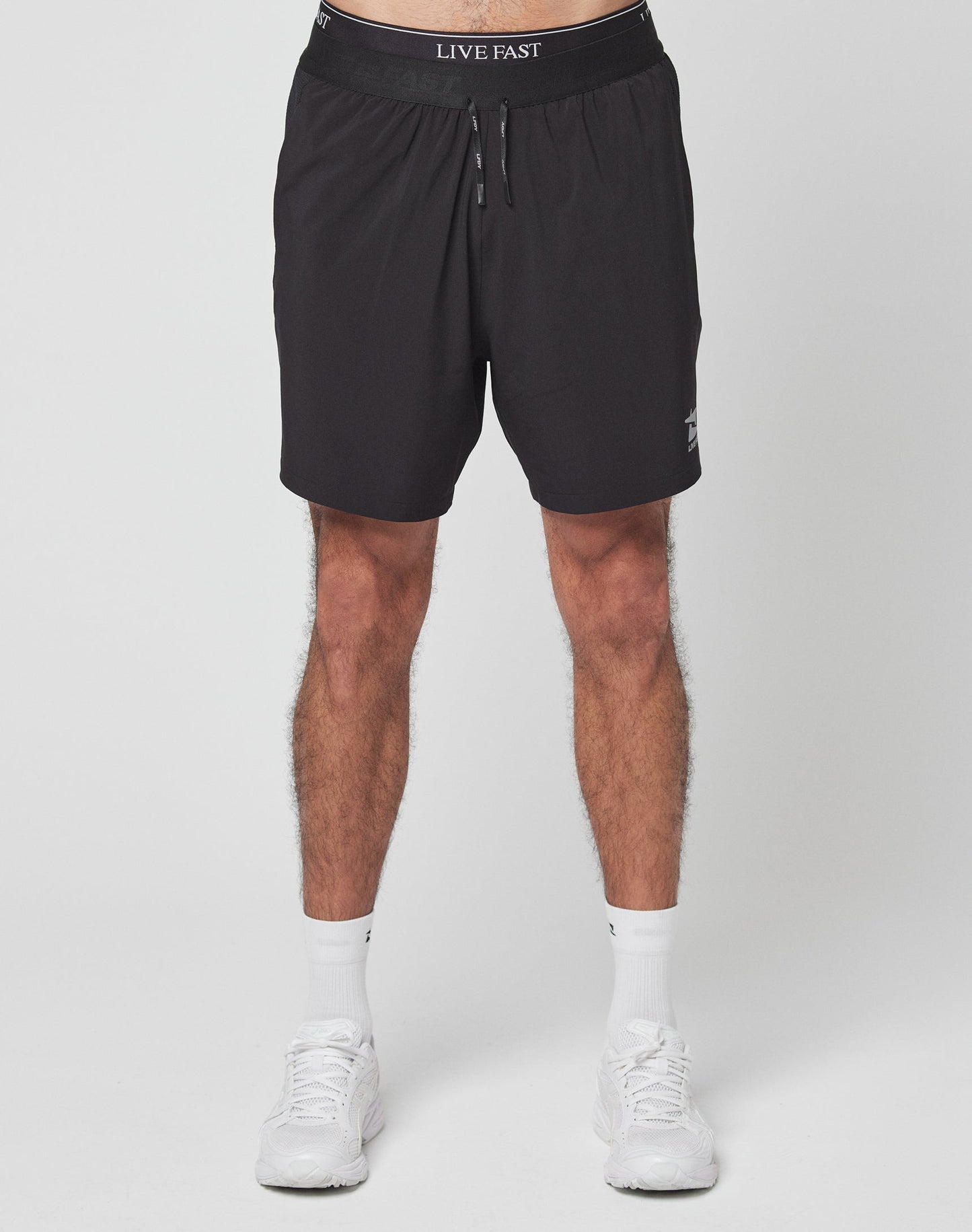 A person stands against a plain white background wearing LIVE FAST DIE YOUNG’s Live Fast Performance Shorts—black with LIVE FAST on the elastic waistband, regular fit—paired with white socks and sneakers. The photo is cropped at the waist and ankles.