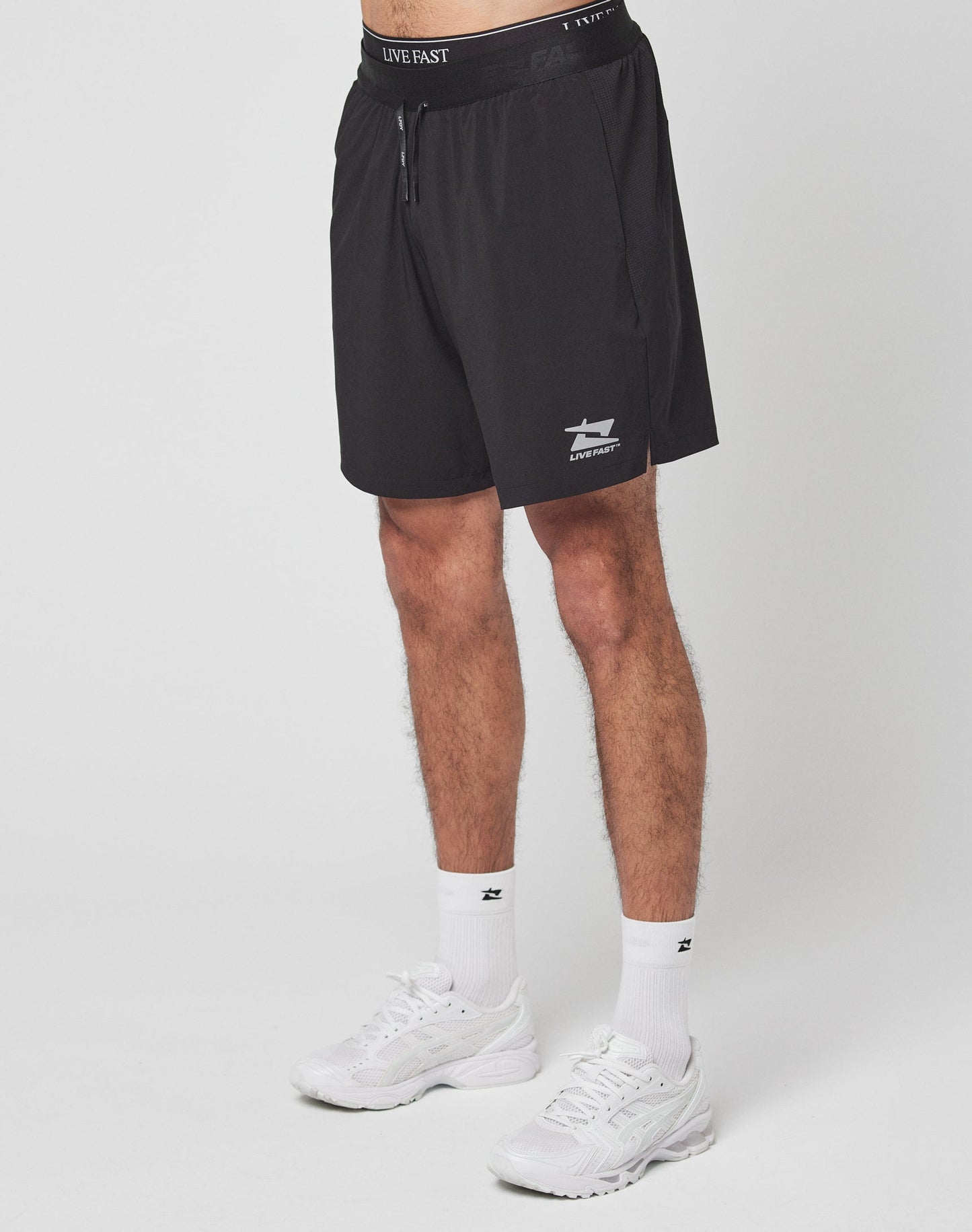 Someone wearing LIVE FAST DIE YOUNG Live Fast Performance Shorts in black—with a shark logo, reflektierender Print, and Elastikbund—plus white socks and sneakers stands against a plain white background. The upper body is not visible.| model