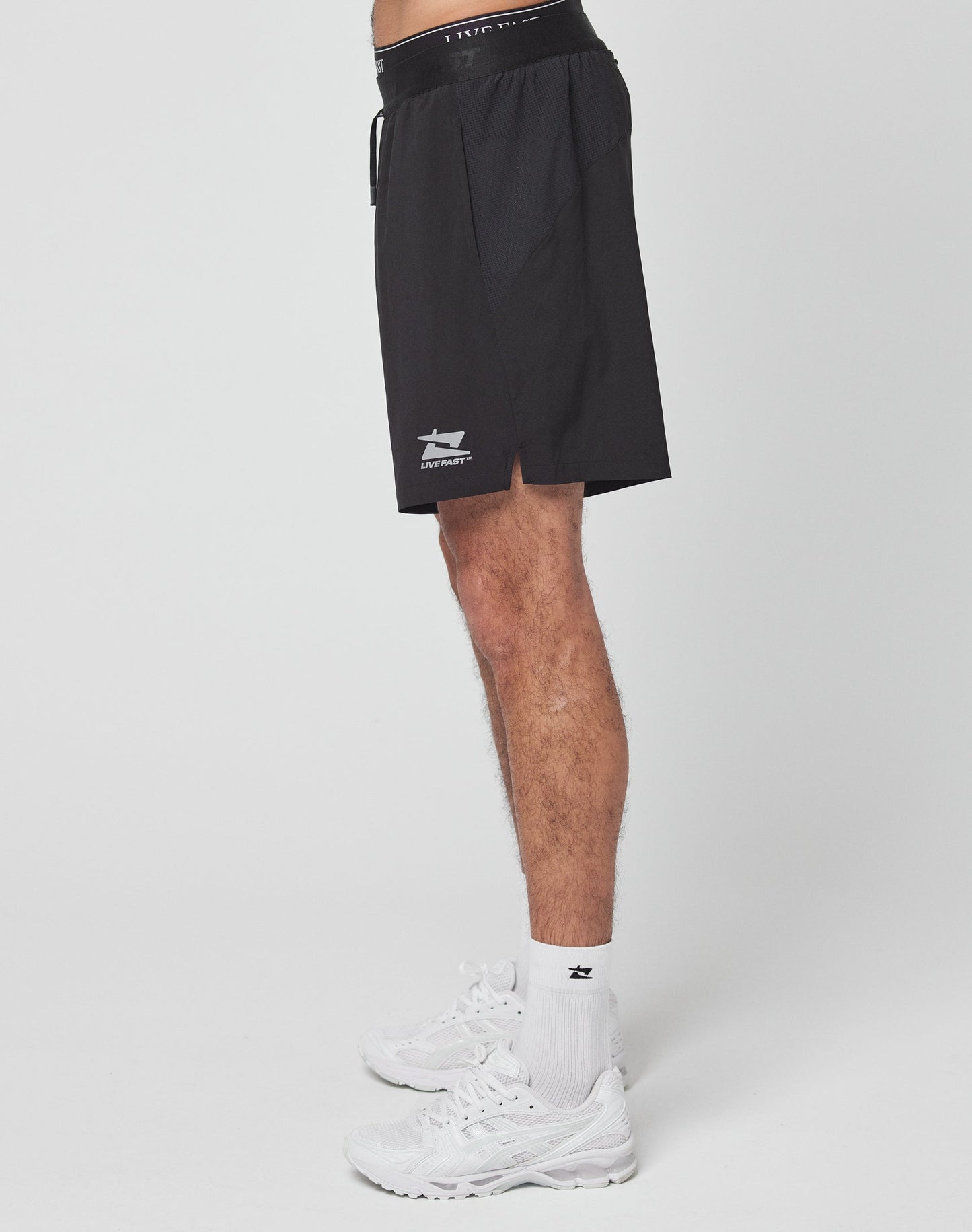 A person stands sideways wearing LIVE FAST DIE YOUNG Live Fast Performance Shorts with elastic waistband and reflective print, paired with white socks and sneakers against a plain white background.