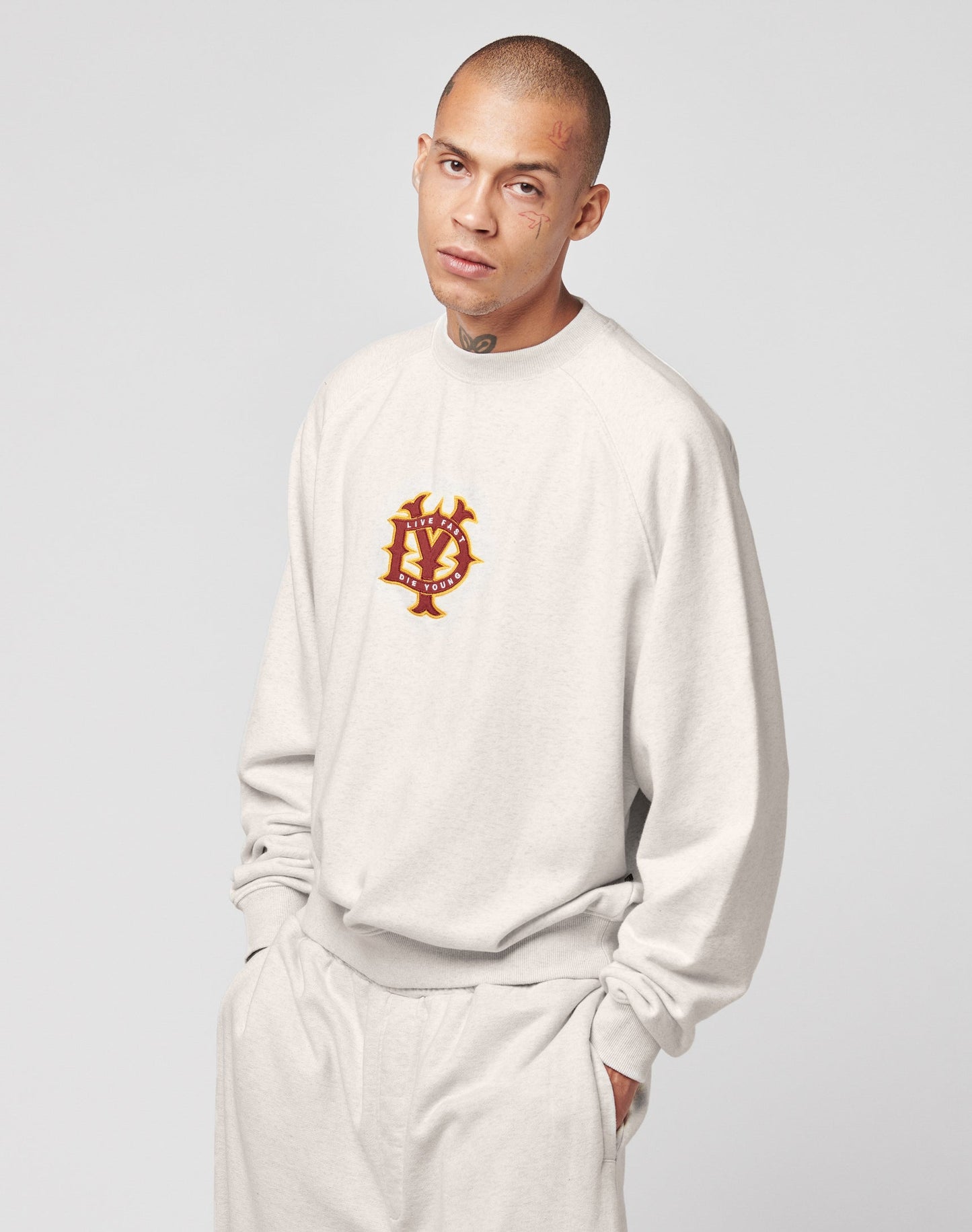 A person with a neutral expression wears the LIVE FAST DIE YOUNG DY Sweater in an oversize fit, light beige color with raglan sleeves and a red and gold crest emblem, paired with matching pants against a plain light gray background.