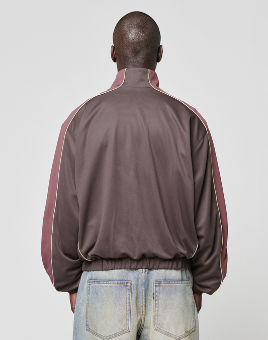 A person faces away, wearing the LIVE FAST DIE YOUNG LFDY x Baba Angora Trackjacket in brown and mauve with white piping, paired with faded blue jeans against a plain light gray background.