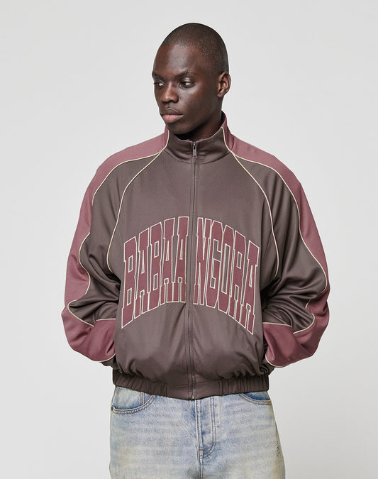 A person wears the LFDY x Baba Angora Trackjacket by LIVE FAST DIE YOUNG, an oversized brown and maroon zip-up with "BABA NGOMA" on the front, standing against a plain background and looking to the side with hands in pockets.