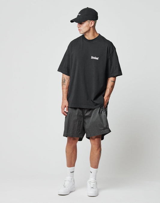 A man in a Regular Fit black cap, loose Tinted Black t-shirt, Live Fast Swimshorts by LIVE FAST DIE YOUNG, white crew socks, and white sneakers stands against a plain white background, looking right with hands in pockets.