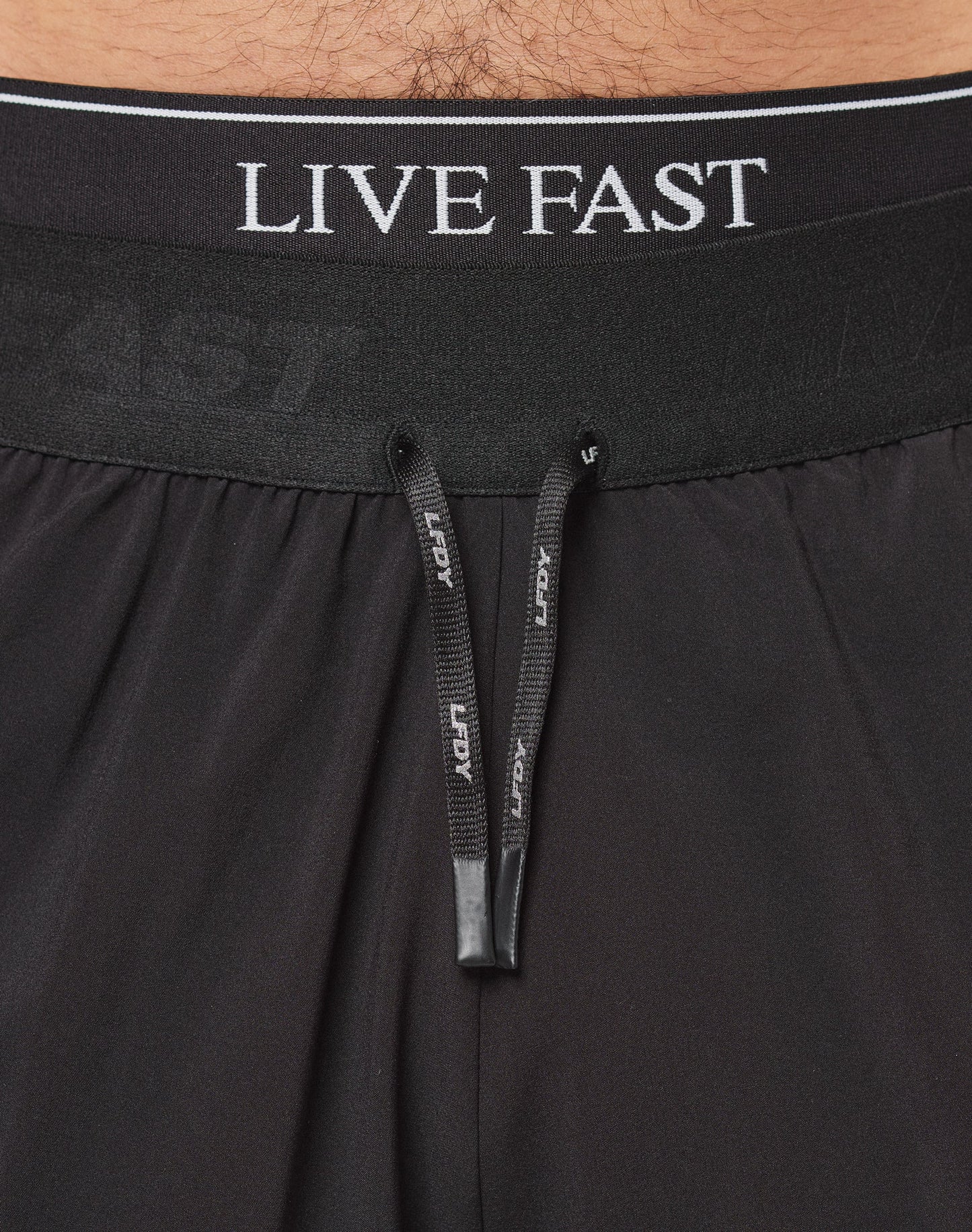 Someone wearing LIVE FAST DIE YOUNG Live Fast Performance Shorts in black—with a shark logo, reflektierender Print, and Elastikbund—plus white socks and sneakers stands against a plain white background. The upper body is not visible.| model