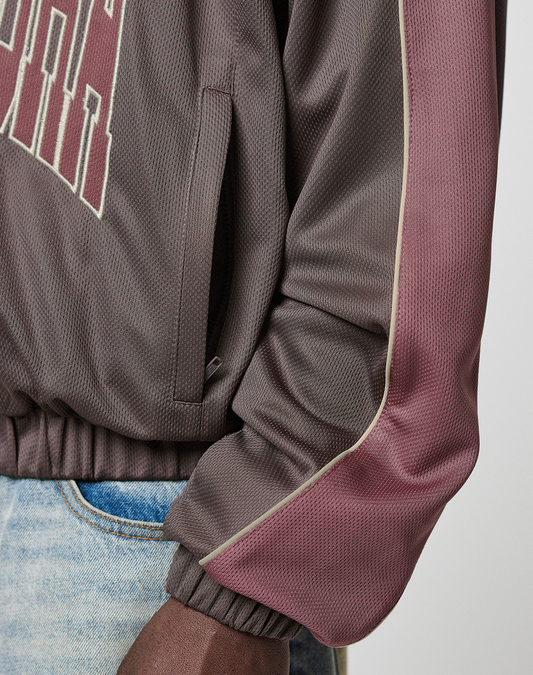 A close-up of a person wearing the LFDY x Baba Angora Trackjacket by LIVE FAST DIE YOUNG, an oversize brown and maroon jacket with lettering, beige piping on the sleeve, paired with light blue jeans and their hand by their side.