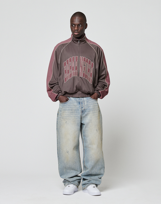 A person stands against a plain background wearing the LIVE FAST DIE YOUNG LFDY x Baba Angora Trackjacket, loose brown with bold Stick Applikation, paired with light blue oversized baggy jeans and white sneakers.