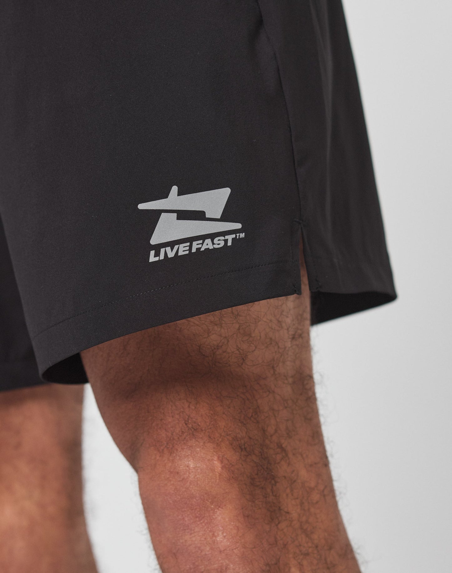 Close-up of a person wearing LIVE FAST DIE YOUNGs Live Fast Performance Shorts. The black Regular Fit shorts feature an elastic waistband, reflective logo print, and LIVE FAST text on the lower left leg.
