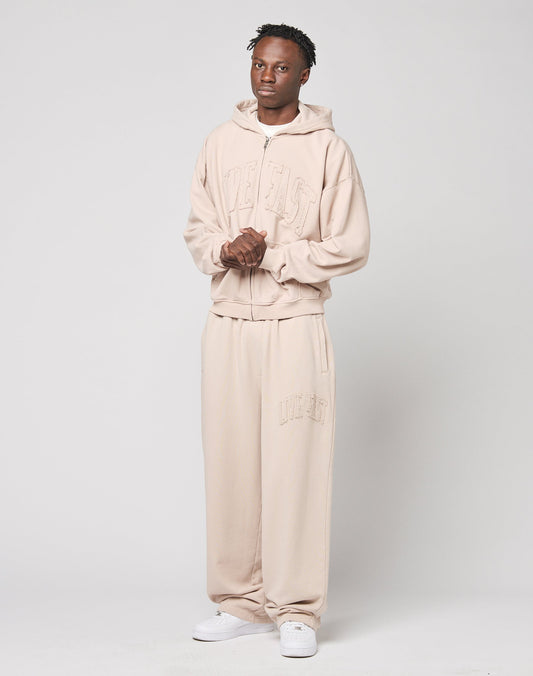 A person stands against a plain background wearing LIVE FAST DIE YOUNG’s Ripped College Wide Jogger in light beige cotton, paired with a matching wide hoodie and white sneakers. Embossed text adds detail as their hands are clasped in front.