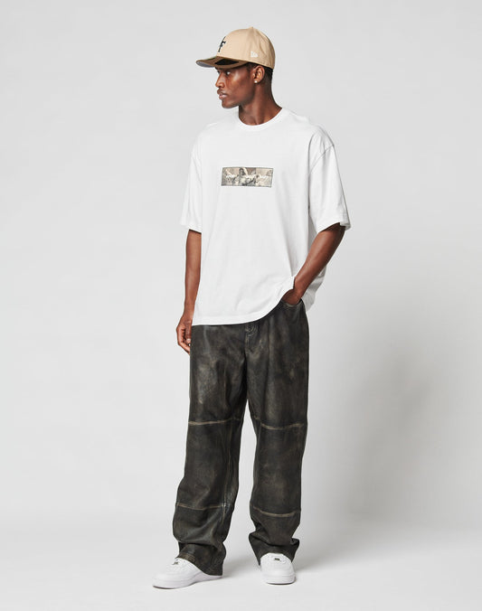 A man in a beige cap, white LFDY Trinity Tee by LIVE FAST DIE YOUNG with reflective print, loose dark pants, and white sneakers stands against a plain light background, looking to the side with one hand in his pocket.
