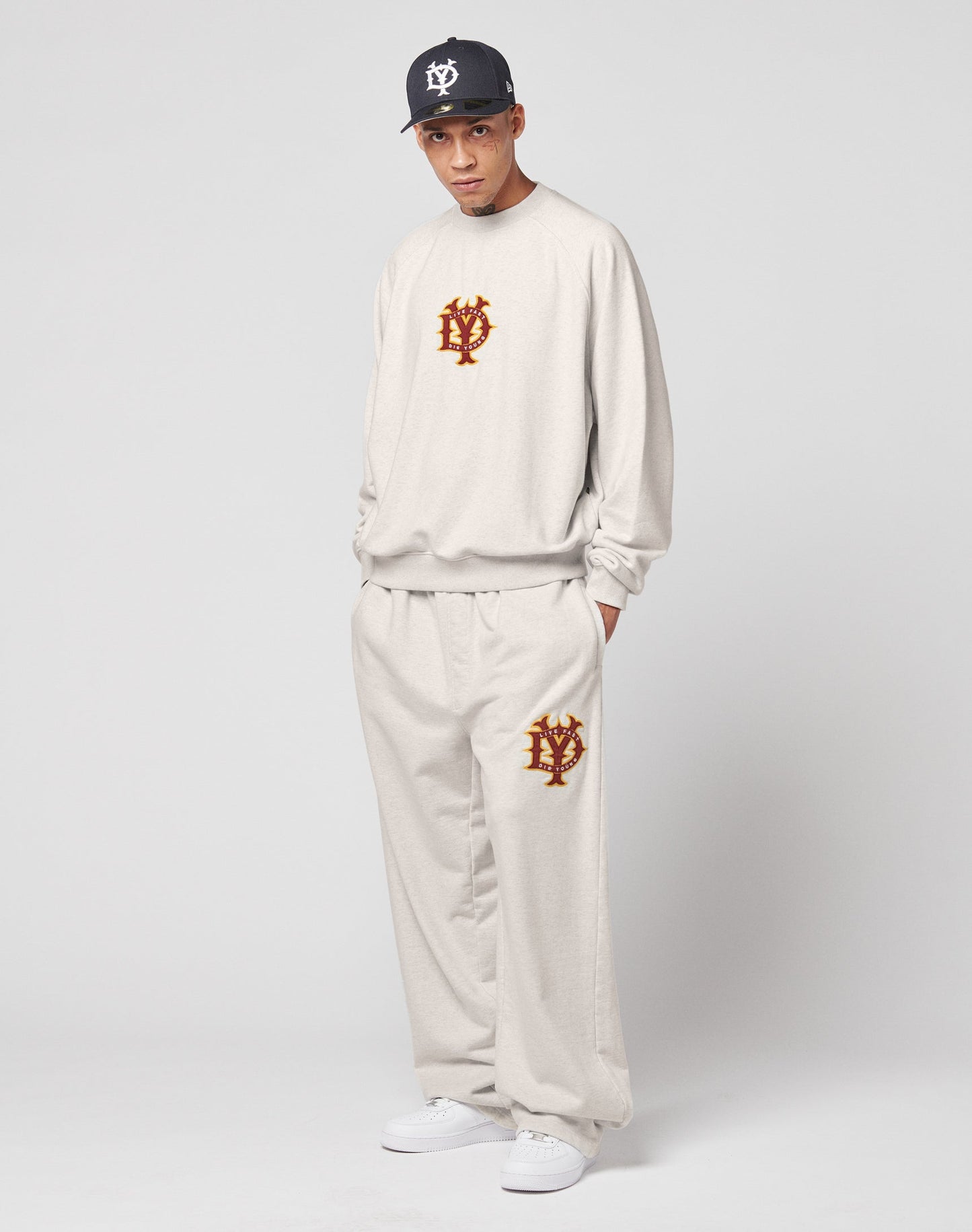 A man in a LIVE FAST DIE YOUNG DY Jogger set—an oversize fit beige sweatshirt and matching wide-leg sweatpants with red and gold NY logos—stands against a plain backdrop, styled with a black cap and white sneakers. Made from 100% cotton.