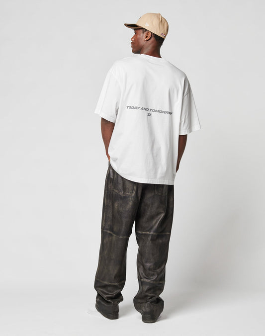 A person faces away, wearing a beige cap, loose dark textured pants (100% Baumwolle), and the LIVE FAST DIE YOUNG LFDY Trinity Tee—an oversized white T-shirt with TODAY AND TOMORROW reflective print—against a plain light background.