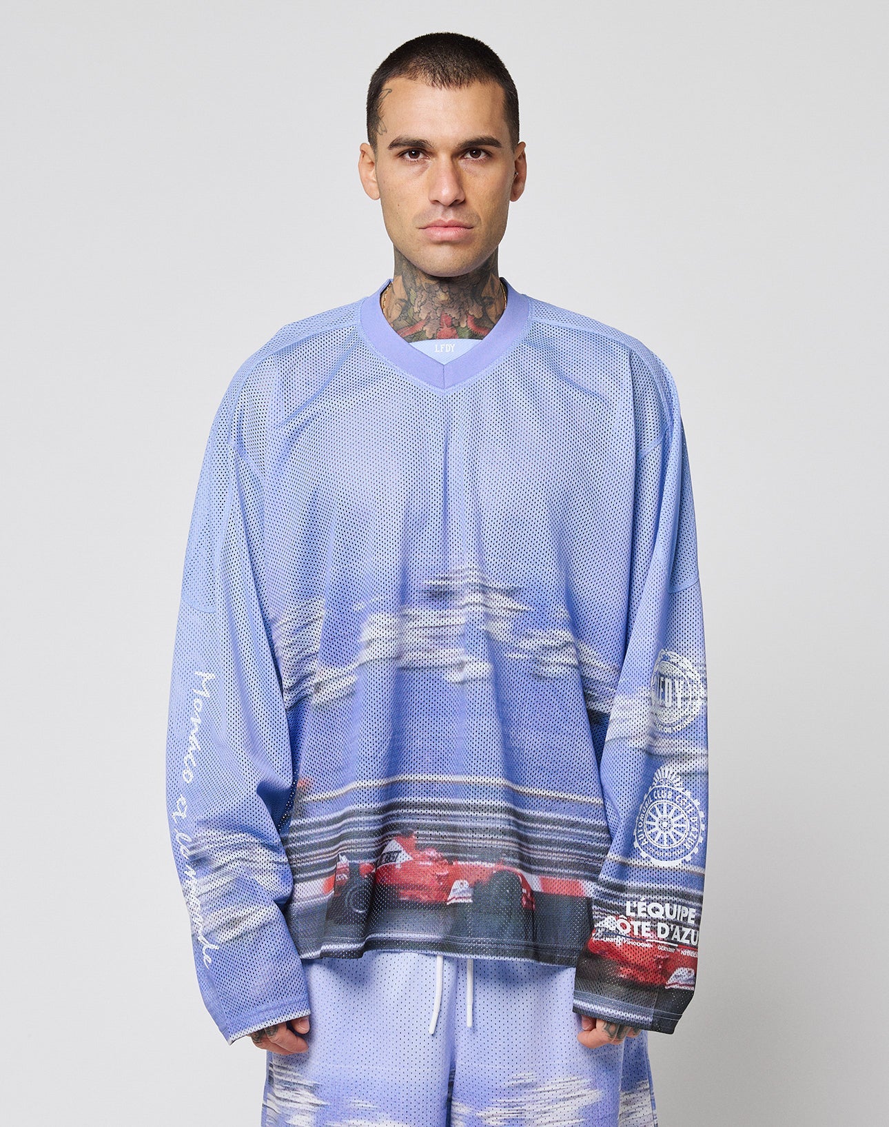 A man with short hair and neck tattoos wears the LIVE FAST DIE YOUNG LFDY Yacht Jersey—an oversize, long-sleeve mesh top with matching shorts in a blue and purple racing car print, set against a plain light background.
