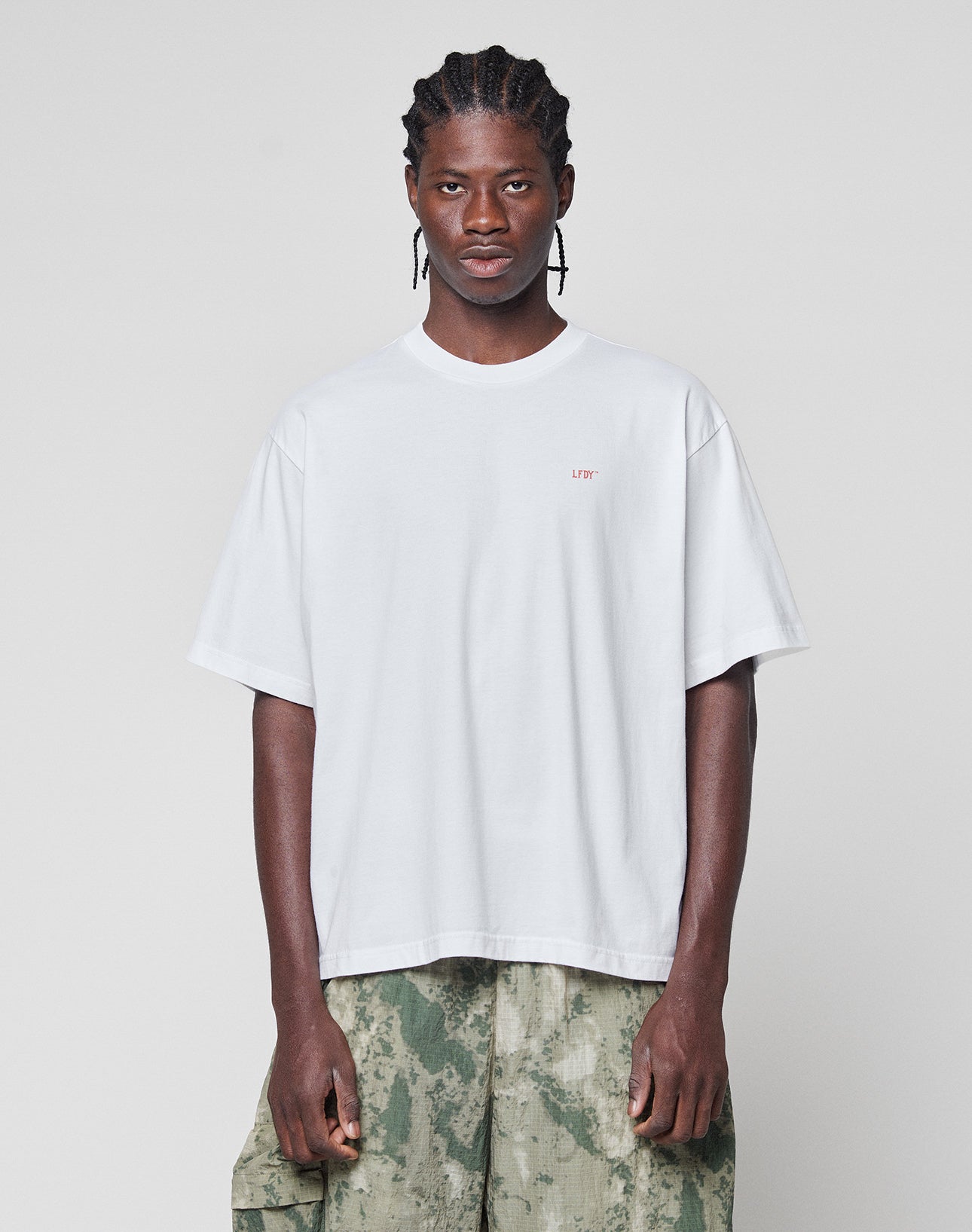 A person with braided hair wears the LIVE FAST DIE YOUNG Live Fast Vegas Tee, a loose white T-shirt with a small red logo on the left chest, and green camo cargo shorts, standing against a plain light gray background.