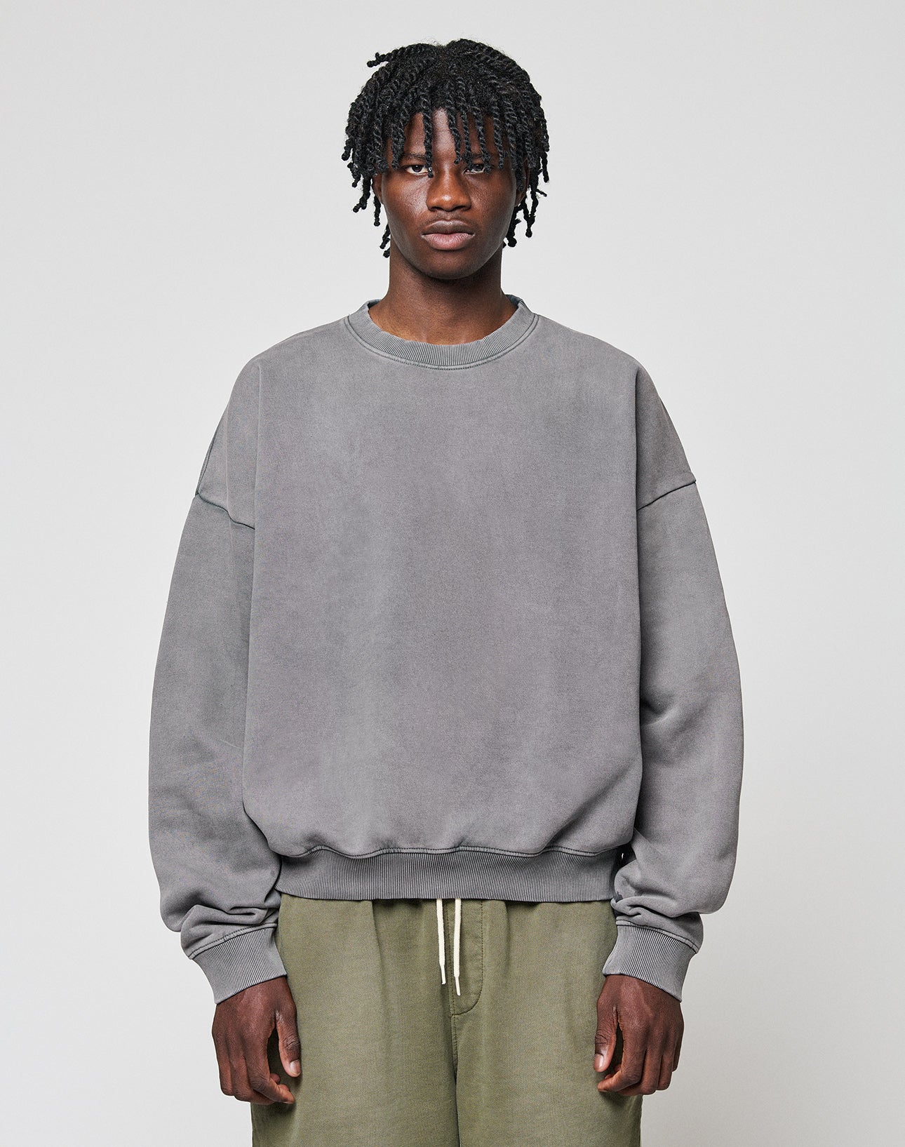 A person with short, twisted hair wears a loose grey LIVE FAST DIE YOUNG Basic Blank Sweater and olive green pants, standing against a plain light background and looking directly at the camera with a neutral expression. | model