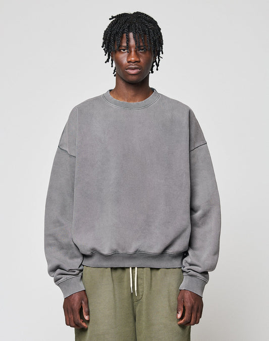 A person with short, twisted hair wears a loose grey LIVE FAST DIE YOUNG Basic Blank Sweater and olive green pants, standing against a plain light background and looking directly at the camera with a neutral expression. | model