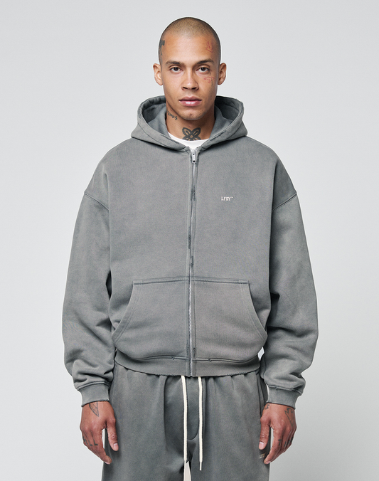 A person in an oversize fit gray Basic Hooded Zip hoodie by LIVE FAST DIE YOUNG, featuring a small white chest logo, front pockets, and matching sweatpants, stands facing forward against a plain light gray background. | model