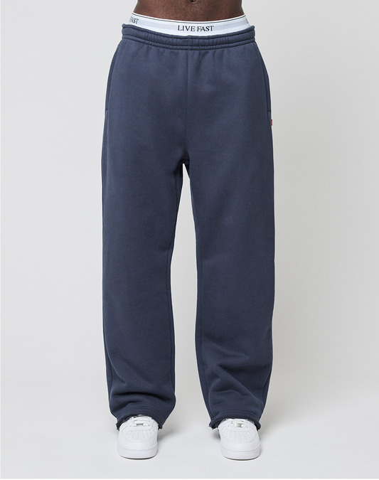 Someone stands against a plain light gray background wearing LIVE FAST DIE YOUNG LFDY Heavyweight Loungepants in navy blue 650gsm, wide fit, with a LIVE FAST waistband and white sneakers.