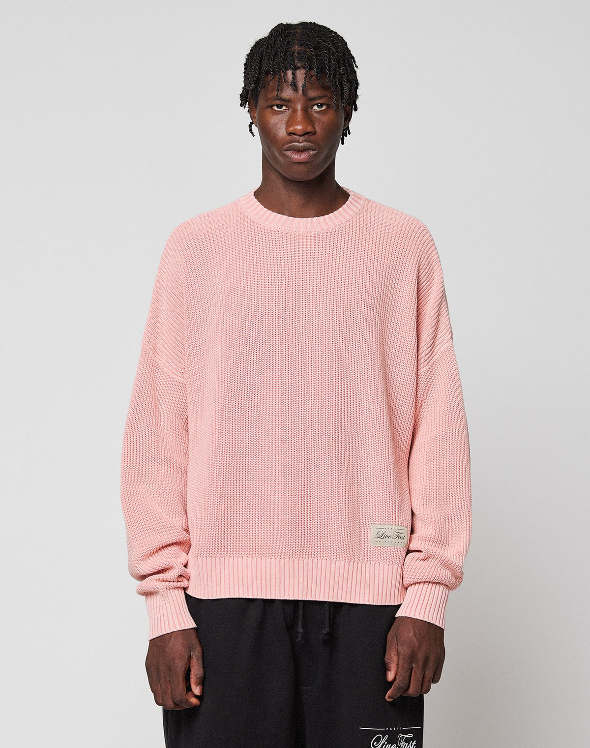 A person with short, textured hair wears the LIVE FAST DIE YOUNG LFDY Knit Sweater in light pink with dropped shoulders and black pants, standing against a plain light gray background. | model