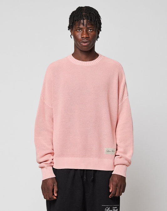 A person with short, textured hair wears the LIVE FAST DIE YOUNG LFDY Knit Sweater in light pink with dropped shoulders and black pants, standing against a plain light gray background. | model