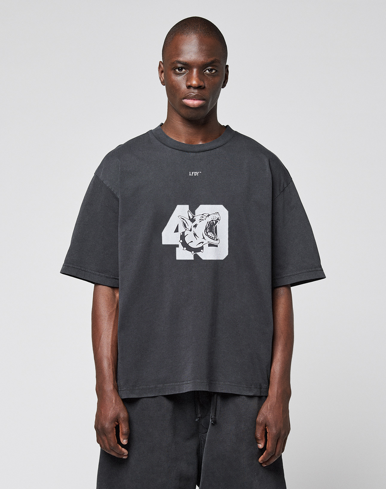 A man faces forward wearing the LIVE FAST DIE YOUNG LFDY 40 Tee—an oversized black T-shirt made of 100% Baumwolle with a bold front print featuring the number 49 and a tiger graphic—paired with black shorts against a light gray backdrop. | model